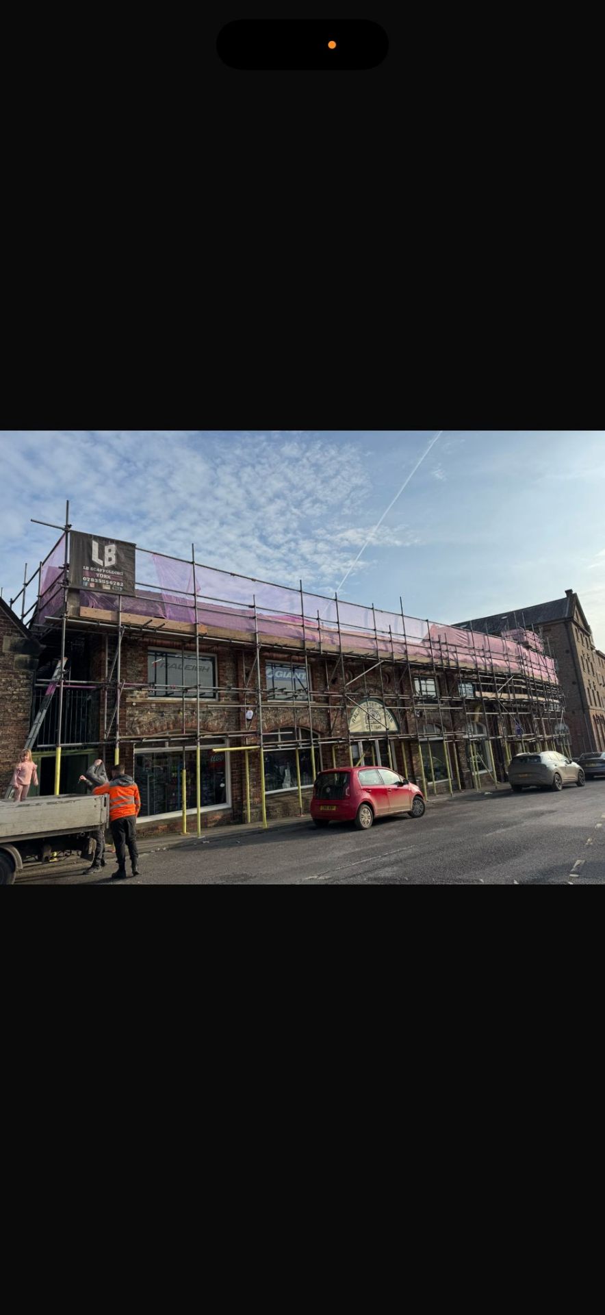 High street shopfront with LB Scaffolding Ltd branded banner and pink safety sheeting along the upper lift